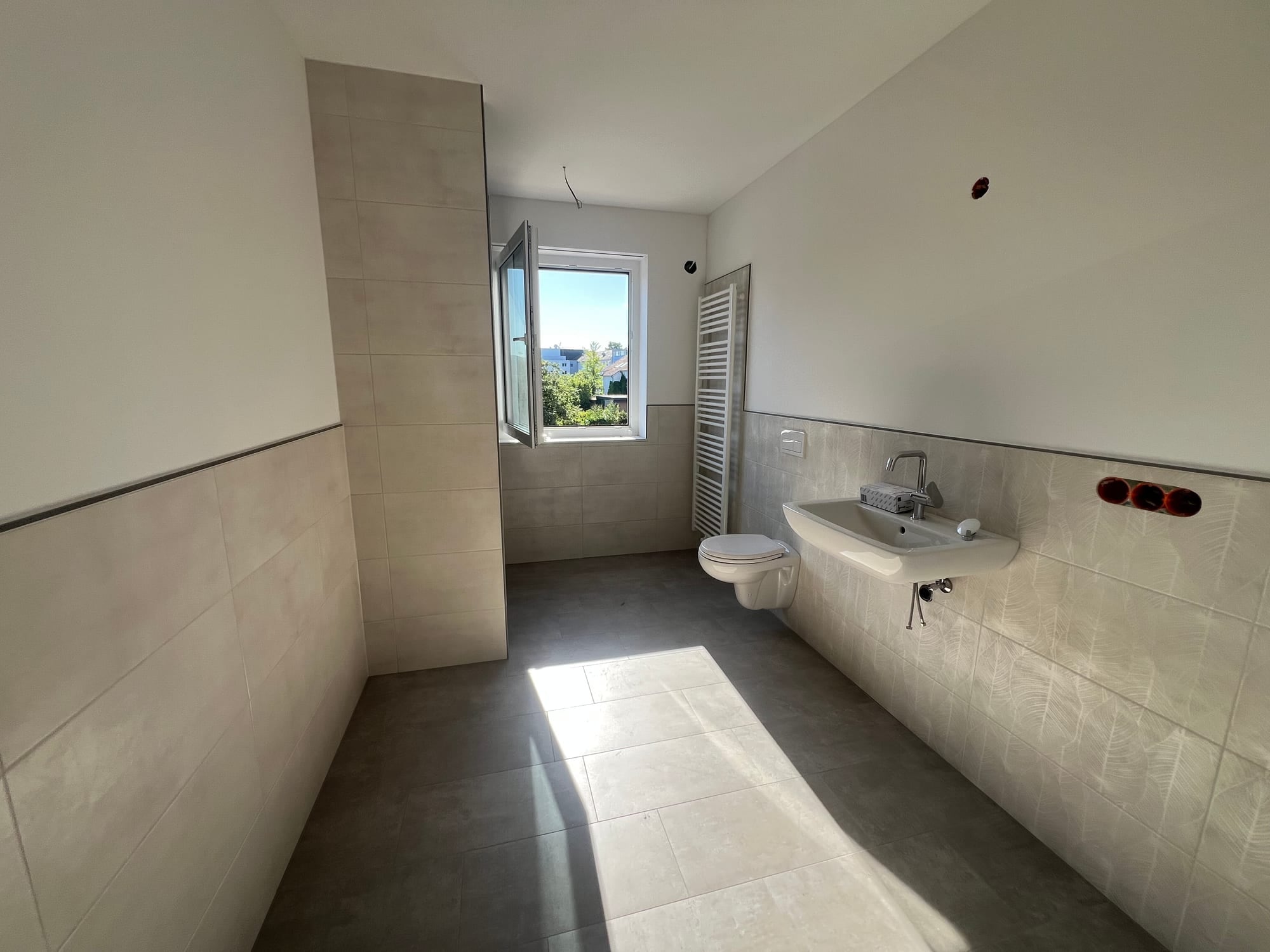 Gefliestes Badezimmer mit bodengleicher Dusche hinter gemauerter Wand, Waschbecken, Wand-WC und Handtuchheizkörper