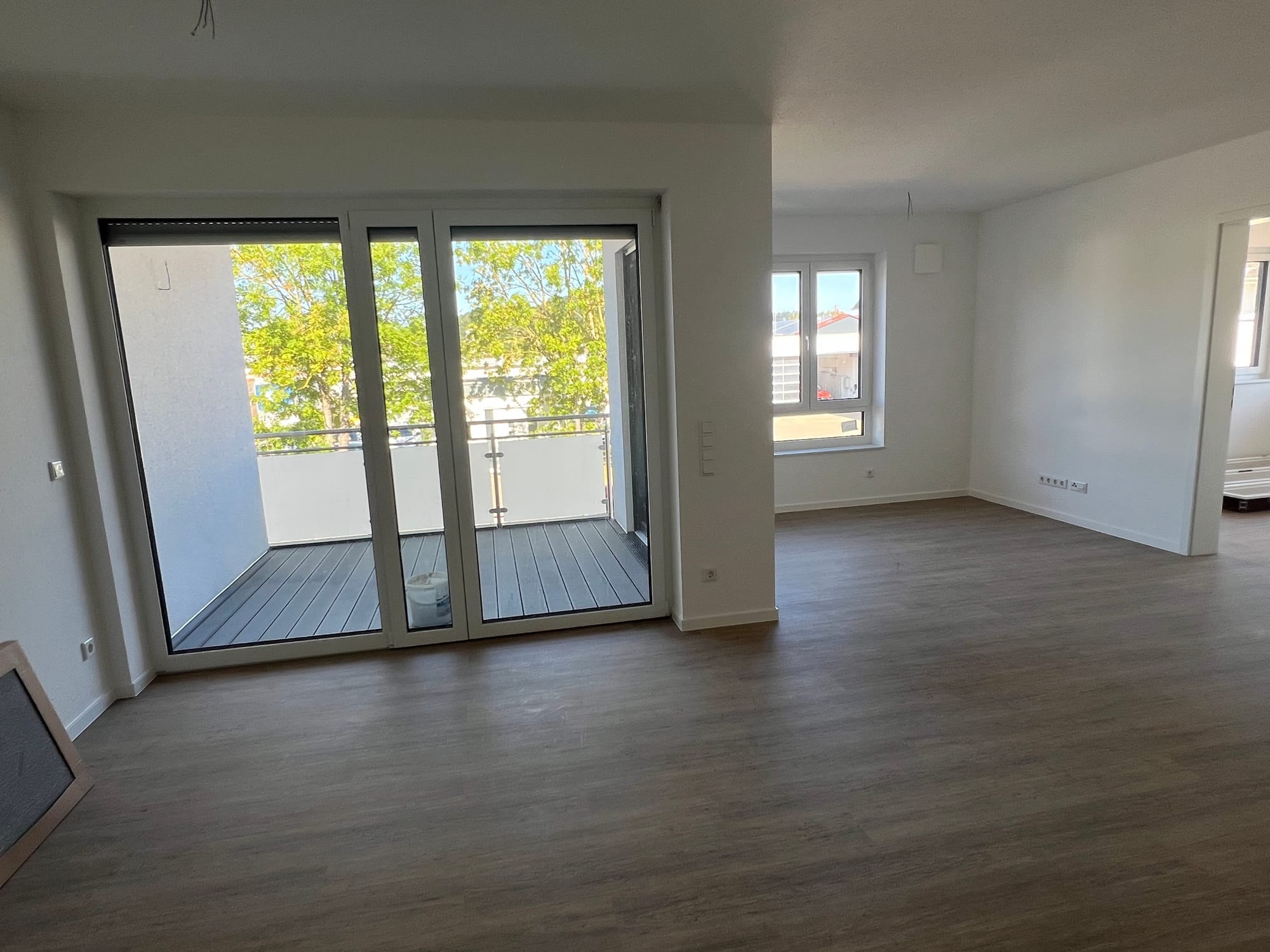 Fertiggestellter Wohnraum mit Vinylboden und bodentiefer Balkontür, dahinter der Balkon mit Holzdielen